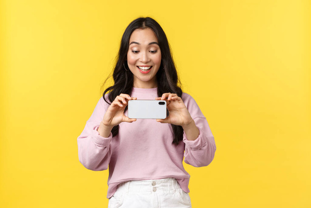 People emotions, lifestyle leisure and beauty concept. Excited cute asian girl with smartphone, record video on phone, looking at display, taking photo of friend, standing yellow background - Φωτογραφία, εικόνα
