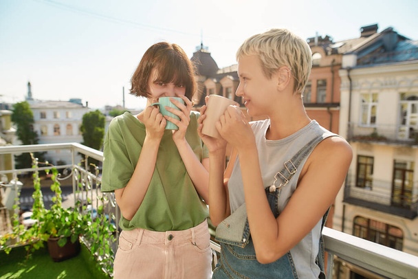 Two cheerful women drinking coffee or tea while standing on the balcony. Young lesbian couple spending their day together - Foto, Imagem