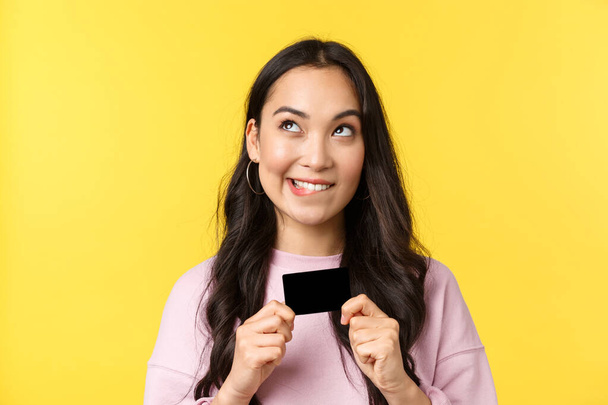 People emotions, lifestyle leisure and beauty concept. Dreamy asian girl thinking what buy next, looking upper left corner and smiling, holding credit card, going online shopping, yellow background - Photo, Image