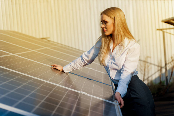 女性は太陽電池パネルにもたれる。ブロンドは発電所で白い正式なシャツを着ていた。家のための無料電気。緑のエネルギー。太陽電池発電事業. - 写真・画像