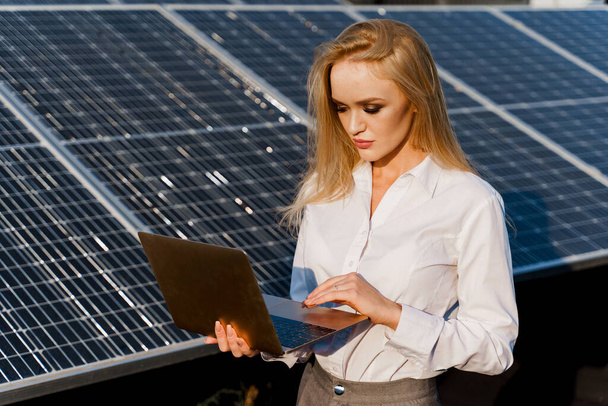 Investor woman stand with laptop near blue solar panels row on the ground Girl weared formal white shirt. Free electricity for home. Sustainability of planet. Green energy. - Foto, imagen