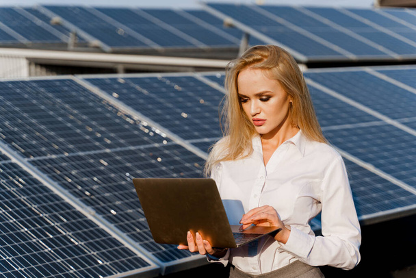 投資家の女性は地面に青い太陽電池パネルの行の近くのノートパソコンでスタンドガールは、正式な白いシャツを着ていた。家のための無料電気。地球の持続可能性。緑のエネルギー. - 写真・画像