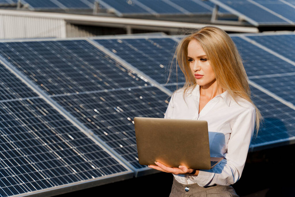 Investor woman stand with laptop near blue solar panels row on the ground Girl weared formal white shirt. Free electricity for home. Sustainability of planet. Green energy. - Φωτογραφία, εικόνα