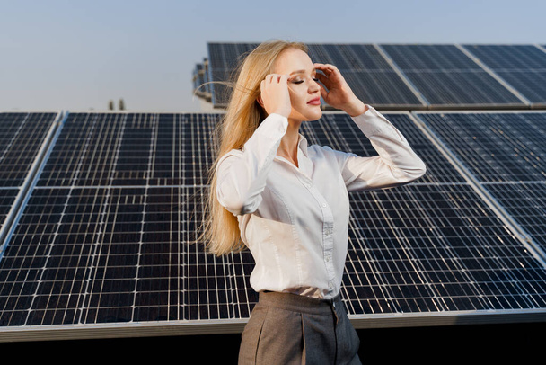 Blonde model with solar panels stands in row on the ground. Girl dressed white formal shirt smiles on the power plant. Free electricity for home. Green energy. Solar cells power plant business. - Фото, изображение