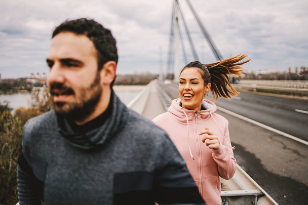 Hymyilevä iloinen urheilullinen pari juoksee sillalla syksyllä pilvisellä säällä ja valmistautuu maratoniin. Terveellisen elämäntavan käsite. Valikoiva keskittyminen nainen. - Valokuva, kuva