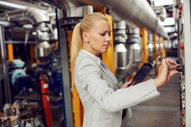 Supervisor femenino rubio dedicado en traje que sostiene la tableta y que se ajusta en el salpicadero mientras está parado en la planta de calefacción. - Foto, imagen