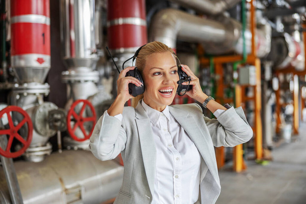 Sonriente mujer exitosa independiente CEO en traje de ponerse auriculares mientras está de pie en su planta de calefacción. - Foto, Imagen