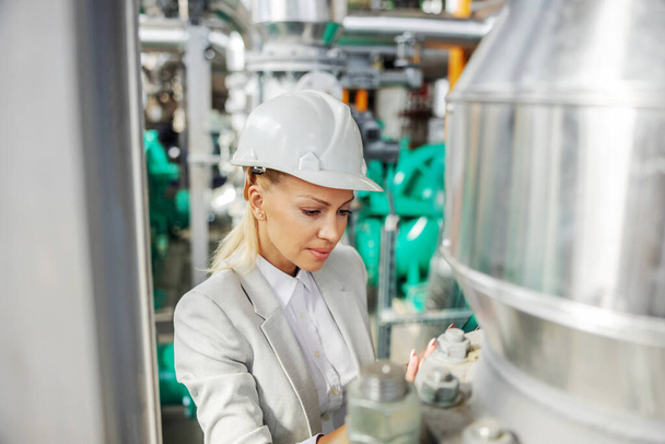 Hardworking dedicated female supervisor in suit with helmet on head standing in heating plant and checking on pressure on turbine. - Photo, Image