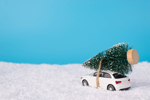 Маленька різдвяна композиція для концепції запрошень. Крупним планом фото маленької іграшкової сучасної машини, що перевозить ялинку на даху, їздить у снігу, подорожуючи
 - Фото, зображення