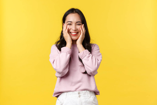 Lidé emoce, životní styl a módní koncept. Rozkošné usmívající se korejská dívka vypadá šťastně, radovat se z velkých úžasných zpráv, držet se za ruce na tváři a stojí žluté pozadí pozitivní - Fotografie, Obrázek
