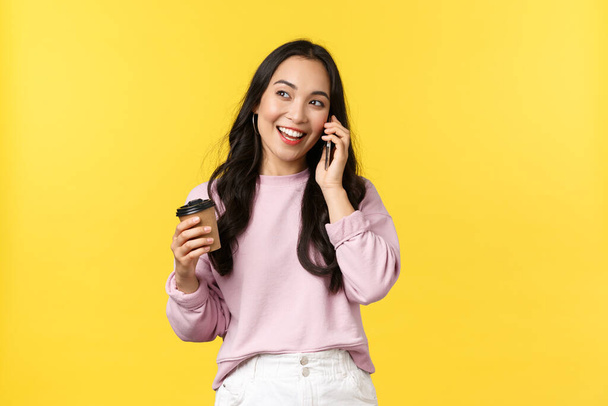 People emotions, lifestyle leisure and beauty concept. Modern cute asian girl talking on phone with friend and smiling, drinking coffee casually, hold takeaway cup, standing yellow background - 写真・画像