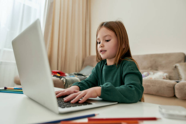 A cute small girl enjoys chatting with her friends via a laptop while sitting alone in a huge bright guestroom alone during distance education - 写真・画像