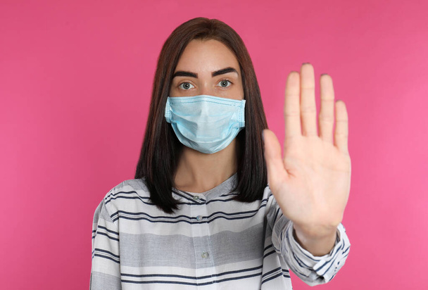 Young woman in protective mask showing stop gesture on pink background. Prevent spreading of coronavirus - Foto, immagini