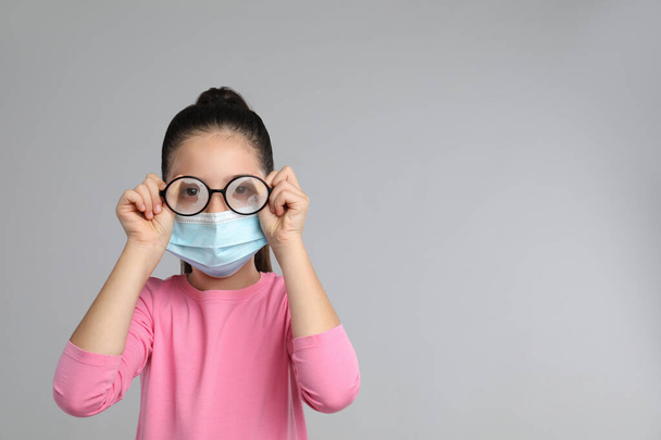 Little girl wiping foggy glasses caused by wearing medical face mask on grey background, space for text. Protective measure during coronavirus pandemic - Foto, afbeelding