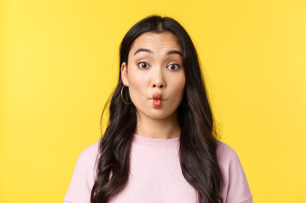 People emotions, lifestyle leisure and beauty concept. Silly and funny asian girl making fish lips and looking indecisive camera, eating something sour, standing yellow background - 写真・画像