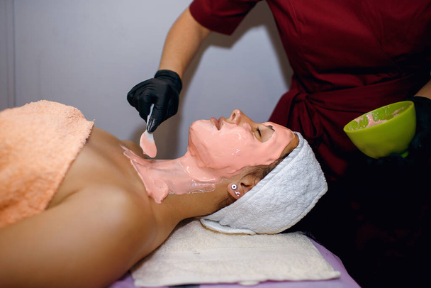 Ein brünettes Mädchen unterzieht sich in einem Wellness-Salon Eingriffen, eine Meisterkosmetikerin in schwarzen Handschuhen trägt eine rosa Maske in einer dicken Schicht auf, reinigt die Haut von Schmutz und überwacht die Reinheit des Gesichts - Foto, Bild