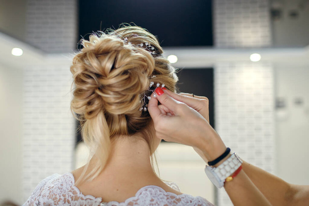 プロの美容師は結婚式の前の朝に若い美しい花嫁に結婚式の髪型を作ります。新郎との会議の準備をしなさい。美しい女性は夫と結婚し - 写真・画像