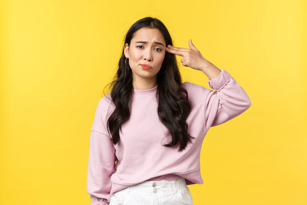 People emotions, lifestyle and fashion concept. Bothered and annoyed asian girl making fake gun over temple as if shooting herself from irritation or distress, standing gloomy yellow background - Foto, Imagem