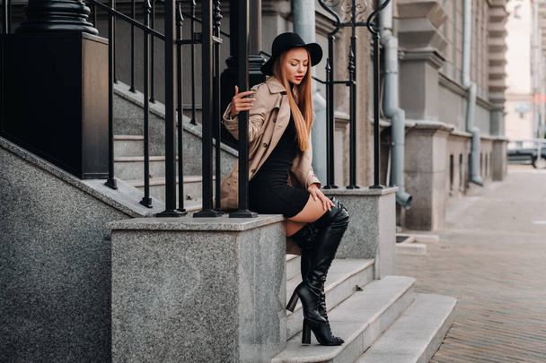 Stylish young woman in a beige coat in a black hat on a city street. Womens street fashion. Autumn clothing.Urban style - Photo, image