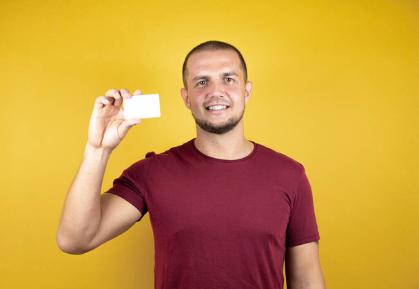 Ruský muž na sobě základní červené tričko přes žluté izolované pozadí zobrazující bílou kartu. - Fotografie, Obrázek