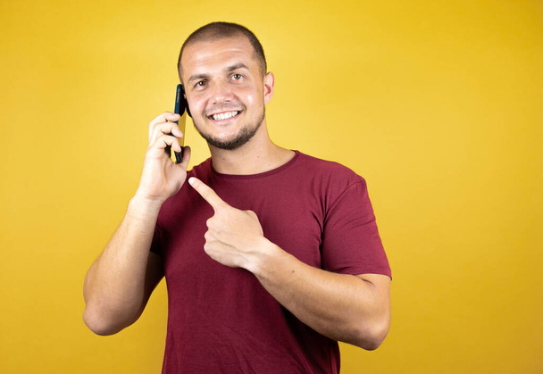 Русский мужчина в обычной красной футболке на желтом инсоляционном фоне разговаривает со смартфоном на жёлтом фоне с удивленным лицом. - Фото, изображение