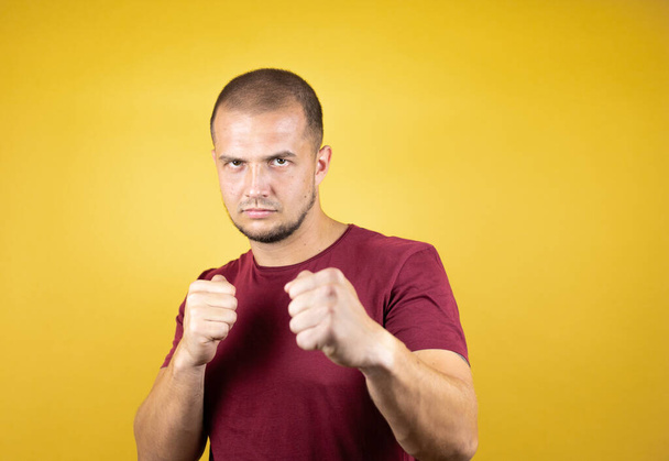 Russian man wearing basic red t-shirt over yellow insolated background Punching fist to fight, aggressive and angry attack, threat and violence - Foto, afbeelding