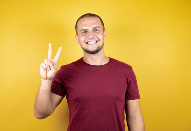Hombre ruso con camiseta roja básica sobre fondo insolado amarillo mostrando y señalando con los dedos número dos mientras sonríe confiado y feliz. - Foto, imagen