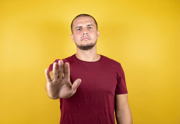 Venäläinen mies yllään perus punainen t-paita yli keltainen eristetty tausta avoimella kädellä tekee stop merkki vakava ja luottavainen ilme, puolustus ele - Valokuva, kuva