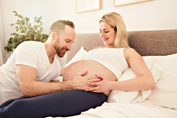 Beautiful pregnant woman and her handsome husband spending time together in bed - 写真・画像