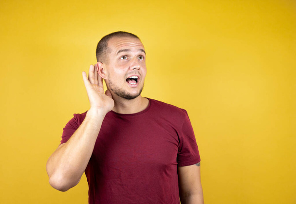 Русский мужчина в обычной красной футболке на желтом инсоляционном фоне улыбается рукой над ухом, слушая слухи или сплетни - Фото, изображение