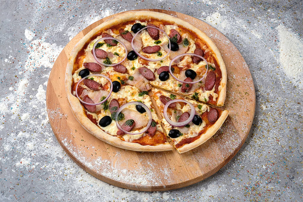 Whole meat Pizza With smoked hunting sausage, Spanish onion rings, black olives and tomato sauce on wooden plate served with culinary flour and spices on gray concrete background. - Photo, Image