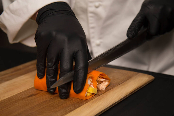 Sushi meester gesneden Japanse geen rijst roll met mes op houten plank. Chef bereidt voedsel in zwarte beschermende handschoenen. Sushi zonder rijst gewikkeld in wortel daikon. Professioneel kookproces  - Foto, afbeelding