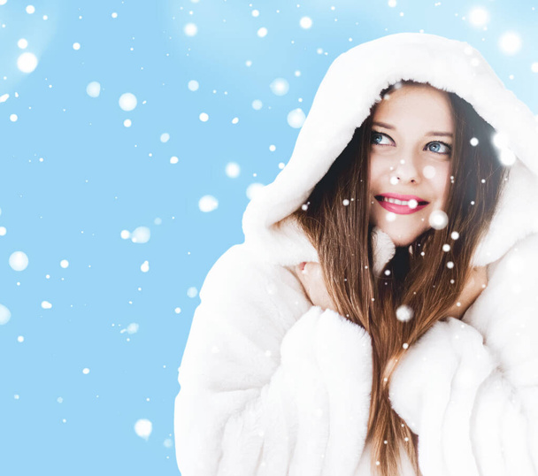 Christmas and winter holiday portrait of young woman in white hooded fur coat, snow on blue background, fashion and lifestyle - Photo, Image