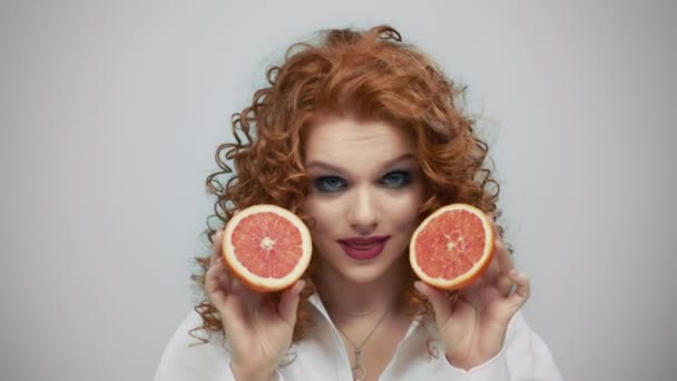 Woman holding grapefruit halves. Cheerful girl biting lips on grey background - Footage, Video