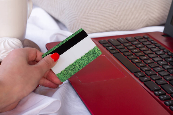 Πιστωτική κάρτα στο χέρι κοντά στο πληκτρολόγιο του ένα κόκκινο φορητό υπολογιστή, στέκεται στο κρεβάτι δίπλα σε ένα σημειωματάριο, μολύβι, κύπελλο, marshmallows και τα cookies. Έννοια online αγορών - Φωτογραφία, εικόνα