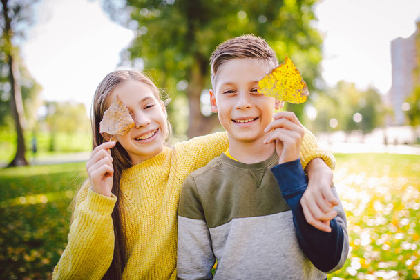 Ευτυχισμένα δίδυμα έφηβοι αγόρι και κορίτσι που ποζάρουν αγκαλιασμένοι στο πάρκο του φθινοπώρου κρατώντας πεσμένα κίτρινα φύλλα στο χέρι σε ηλιόλουστο καιρό. Θέμα φθινοπωρινής περιόδου. Τα αδέρφια διασκεδάζουν παίζοντας με τα φύλλα.. - Φωτογραφία, εικόνα