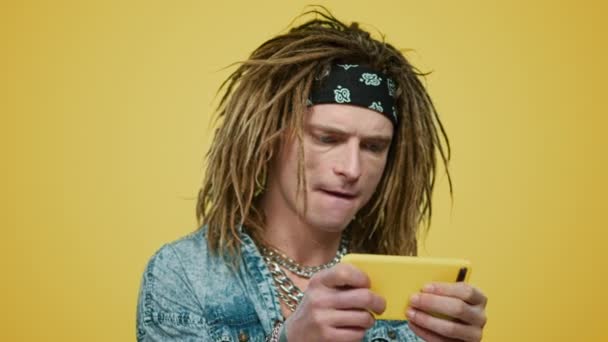 Rocker pelaa peliä älypuhelimella keltaisella taustalla. Tyylikäs kaveri käyttää puhelinta - Materiaali, video