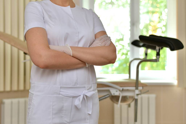Closeup of a doctor in a white uniform, wearing gloves, with folded hands in front of him. Stands in front of a gynecological chair with black footrests. In the medical office. - Foto, imagen