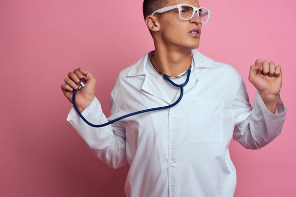 mannelijke arts in een medische jurk met een stethoscoop rond zijn nek op een roze achtergrond en bril op zijn gezicht - Foto, afbeelding