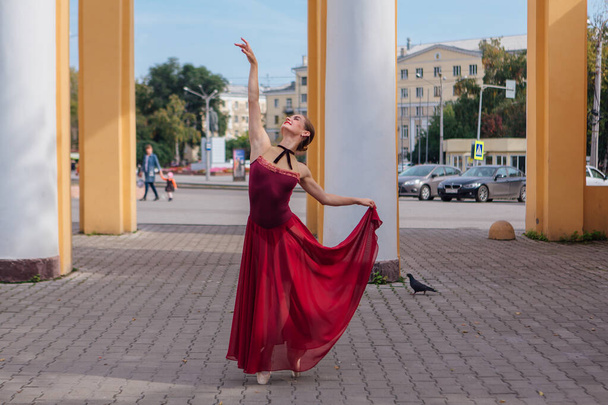 Woman ballerina in red ballet dress dancing in pointe shoes next to the old columns. Ballerina standing in beautiful ballet pose - Fotografie, Obrázek