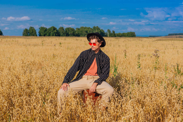 Rural Countryside Scene. Tall handsome man dressed in a black shirt, black hat and pink sunglasses sitting on a brown vintage leather suitcaseat at golden oat field. Summer landscape with blue sky - Фото, изображение