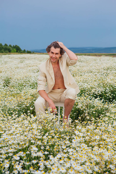 Tall handsome man dressed in a white suit on naked body sitting on a back of a chair in camomile flowers field - Foto, Imagen