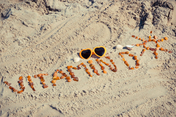 Inscripción vitamina D con forma de sol hecha de piedras de ámbar y gafas de sol sobre arena en la playa. Prevención de la deficiencia de vitamina D. Hora de verano - Foto, imagen