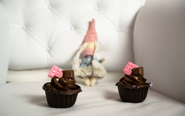 Los cupcakes marrones están decorados con chocolate rosa en un sillón de cuero vintage ligero y una muñeca textil en traje de invierno en el fondo.. - Foto, imagen