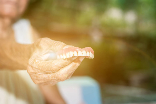古い女性の手は歯ブラスと取り外し可能な部分的な入れ歯の洗浄のために汚れて偽の歯を保持します - 写真・画像
