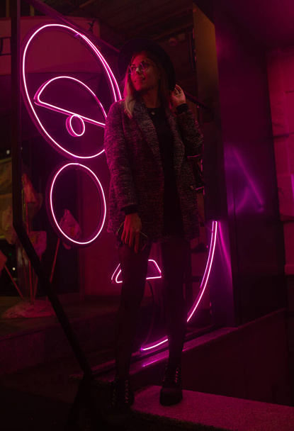 Stylish woman in trendy coat and eyewear looking up enjoying nightlife in city. Gorgeous fashion hipster girl in jacket standing outdoors on street with neon city illumination. - Foto, Imagen