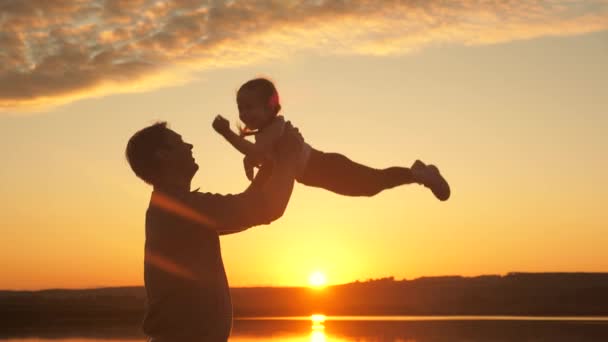 Papà getta la sua figlioletta felice in aria sulla spiaggia, divertendosi al tramonto. padre gioca con il bambino con l'acqua. felice concetto di famiglia. silhouette di un padre e un bambino sano che sorvola il sole - Filmati, video