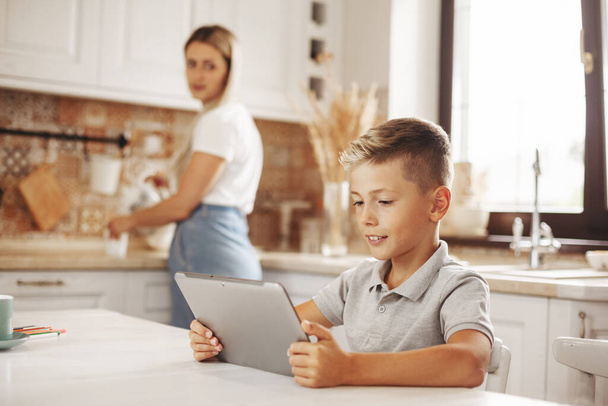 Chłopak odrabia lekcje w kuchni używając tabletu online, rozmawiając z nauczycielem. Mama pilnuje syna. Kształcenie na odległość, edukacja internetowa, koncepcja nauczania w domu - Zdjęcie, obraz