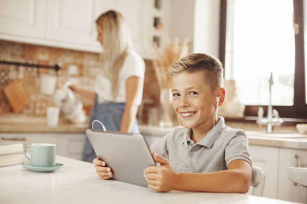 O menino faz seus trabalhos de casa na cozinha no tablet on-line, conversando com o professor. A mãe cozinha e ajuda o filho. Ensino à distância, educação on-line, conceito de escola em casa - Foto, Imagem