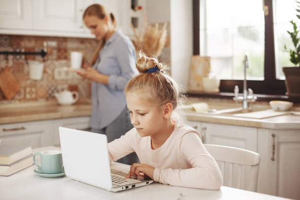 Mom makes tea, and her daughter does her homework in the kitchen with a laptop on the Internet, talking to the teacher. Distance learning, online education, home schooling concept - Photo, Image